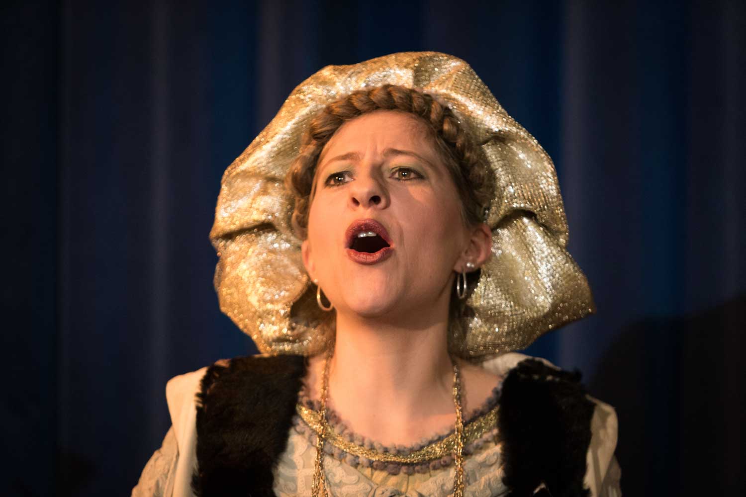 Woman performing in stage costume with a gold headdress, singing passionately against a dark curtain background.