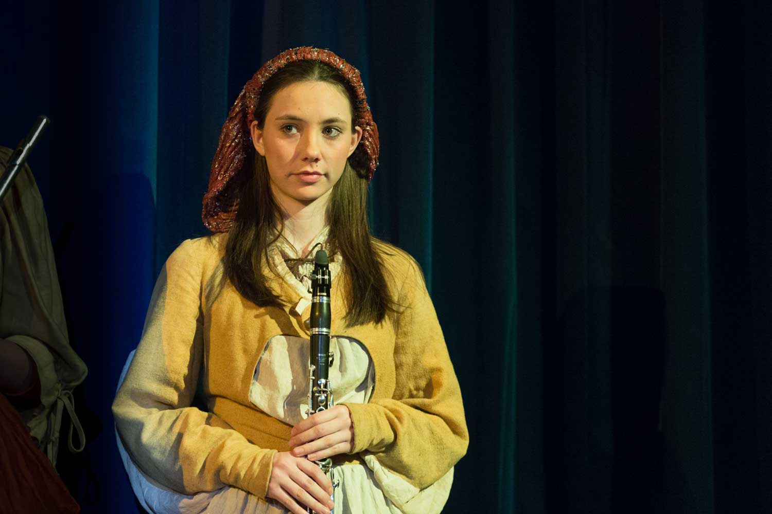 Young woman in medieval costume playing a clarinet on stage, with dark curtain backdrop.