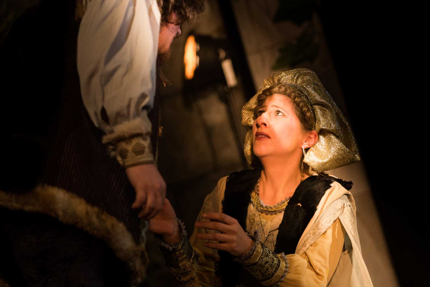 Actor in medieval costume kneels, looking up emotionally at another character, lit by warm torchlight in a dramatic scene.