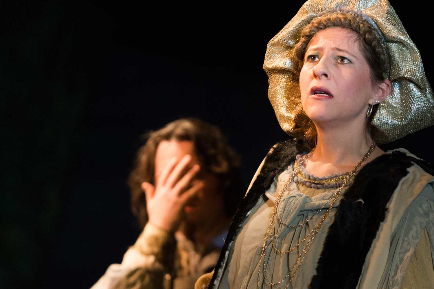 A woman in historical costume looks distressed on stage, while a person in the background covers their face.