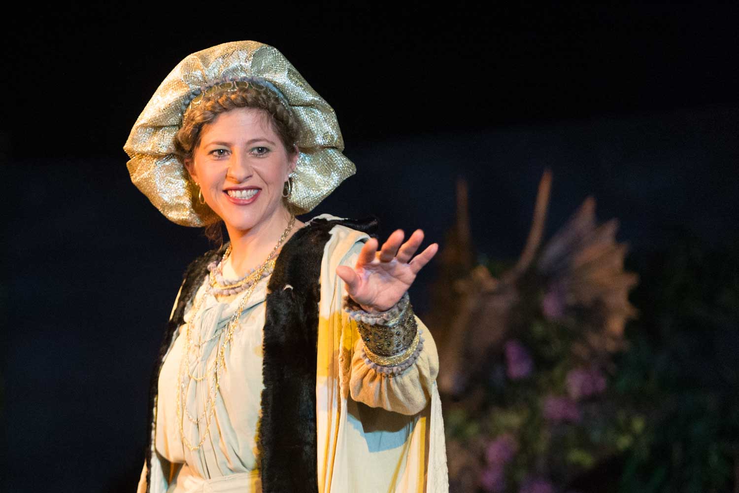 Smiling woman in a medieval costume on stage, reaching out.