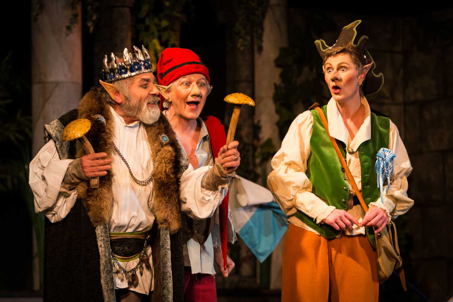 Three actors in fantasy costumes, including a king with a fur cape, holding props on stage during a theatrical performance.