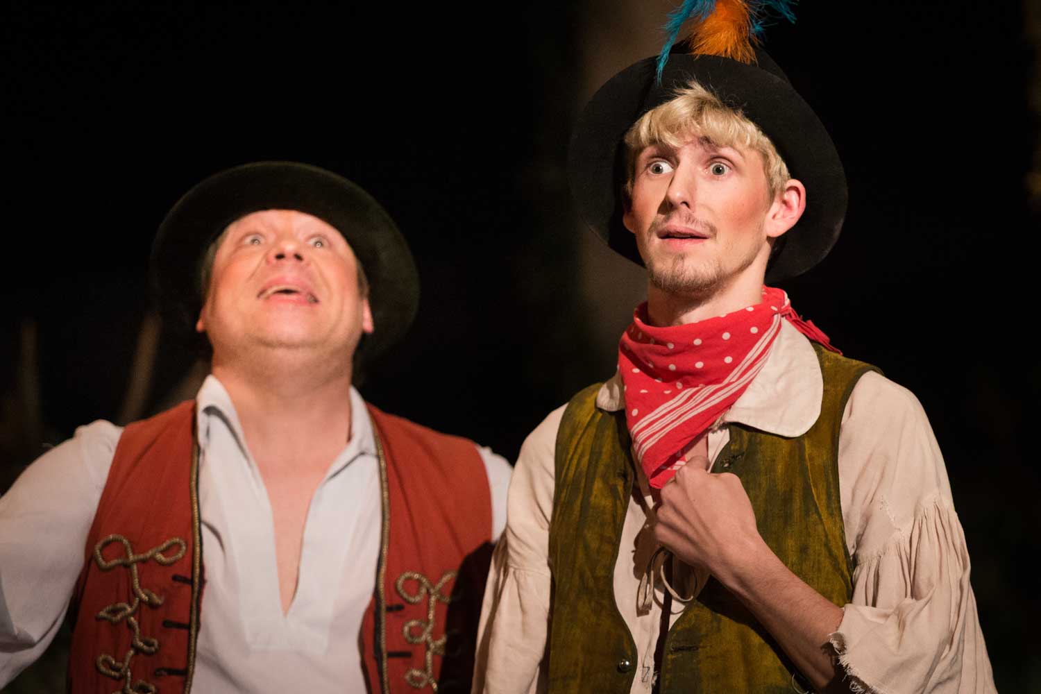 Two actors in vintage costumes, wearing hats and vests, perform expressively on stage with focused expressions.