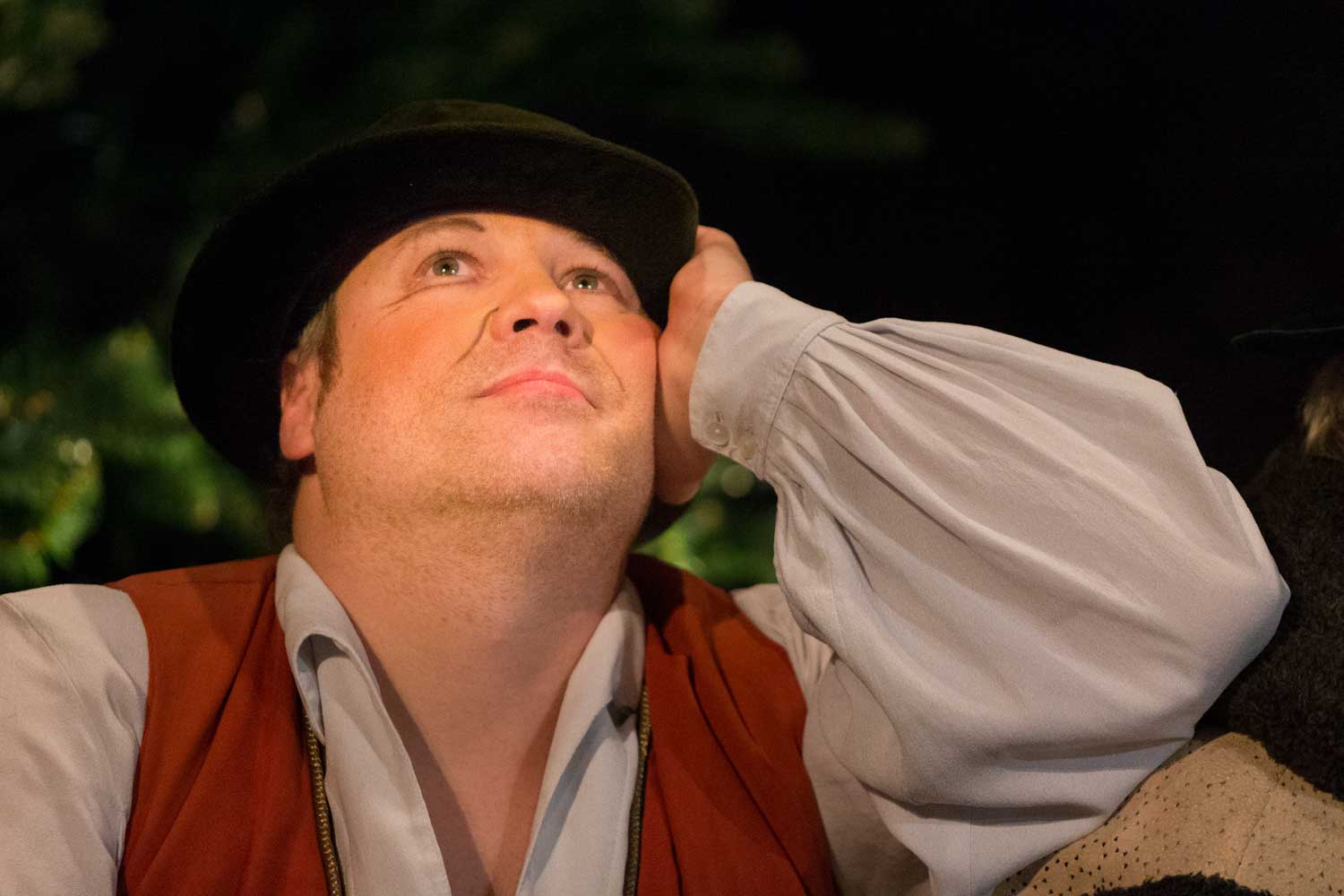 Man in traditional costume with hat gazes thoughtfully upwards, wearing a white shirt and brown vest at a nighttime event.