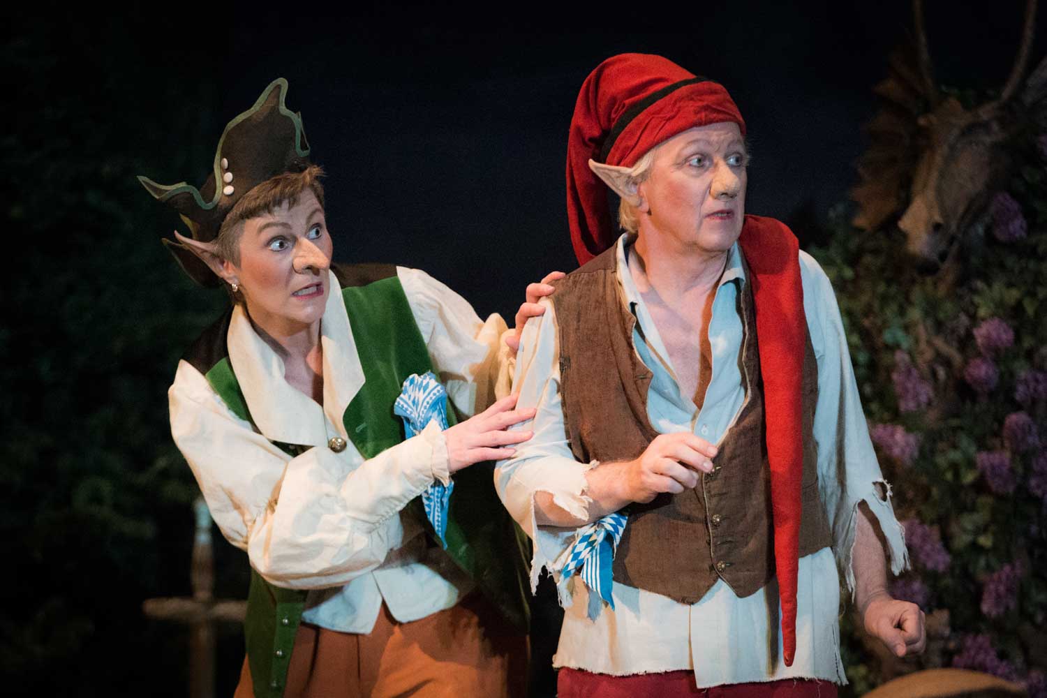 Two actors in elf costumes perform a lively scene on stage, featuring greenery in the background.
