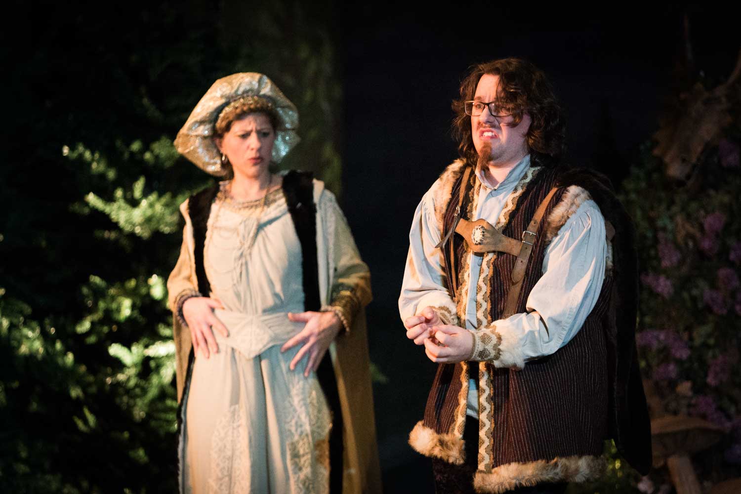 Two actors in medieval costumes performing on stage, surrounded by greenery and soft lighting.