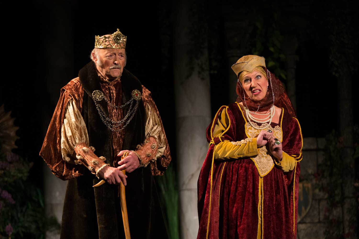 Elderly man in royal costume with crown, holding a cane, stands beside woman in medieval attire on stage.