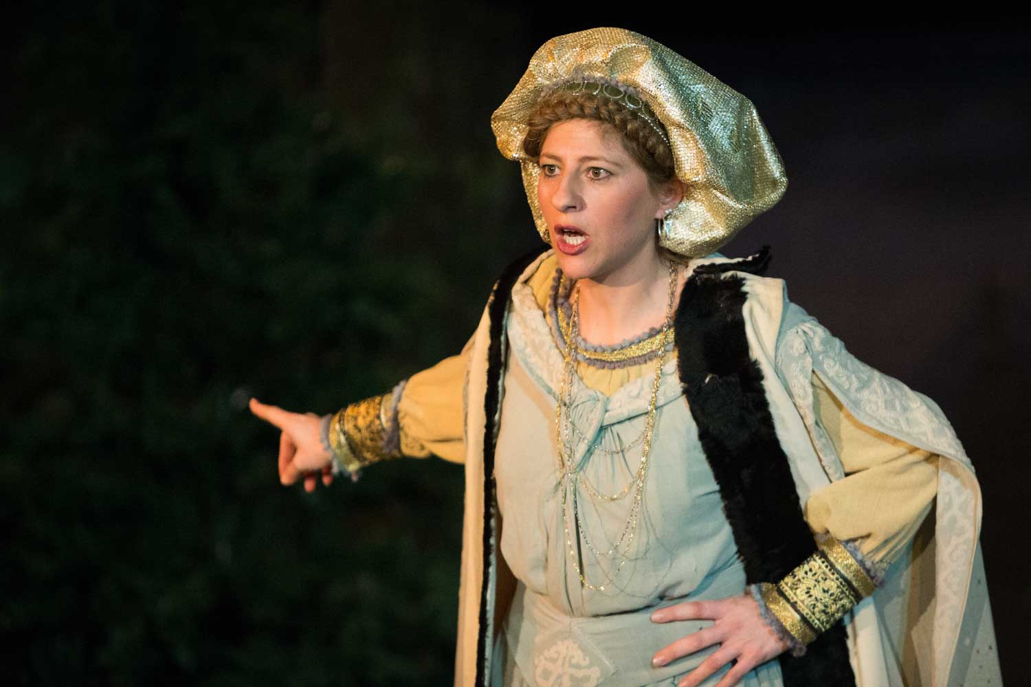 Actress in medieval costume passionately performing on stage, wearing gold headdress and ornate robe against dark backdrop.
