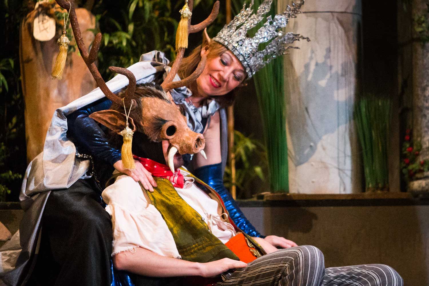 Costumed actors on stage: a queen with a silver crown and a character wearing a boar mask, in a woodland setting.