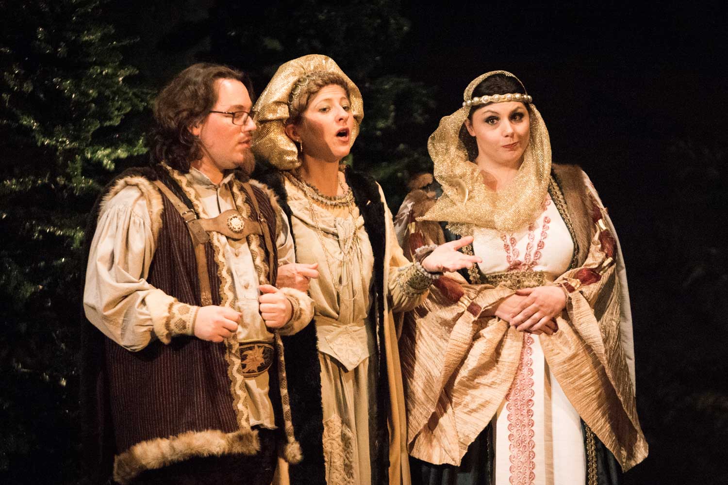 Three performers in medieval costumes sing on stage, with trees in the background.