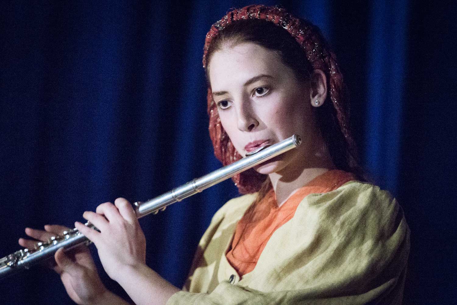 Woman playing flute on stage, wearing a headscarf and colorful attire against a dark blue curtain background.