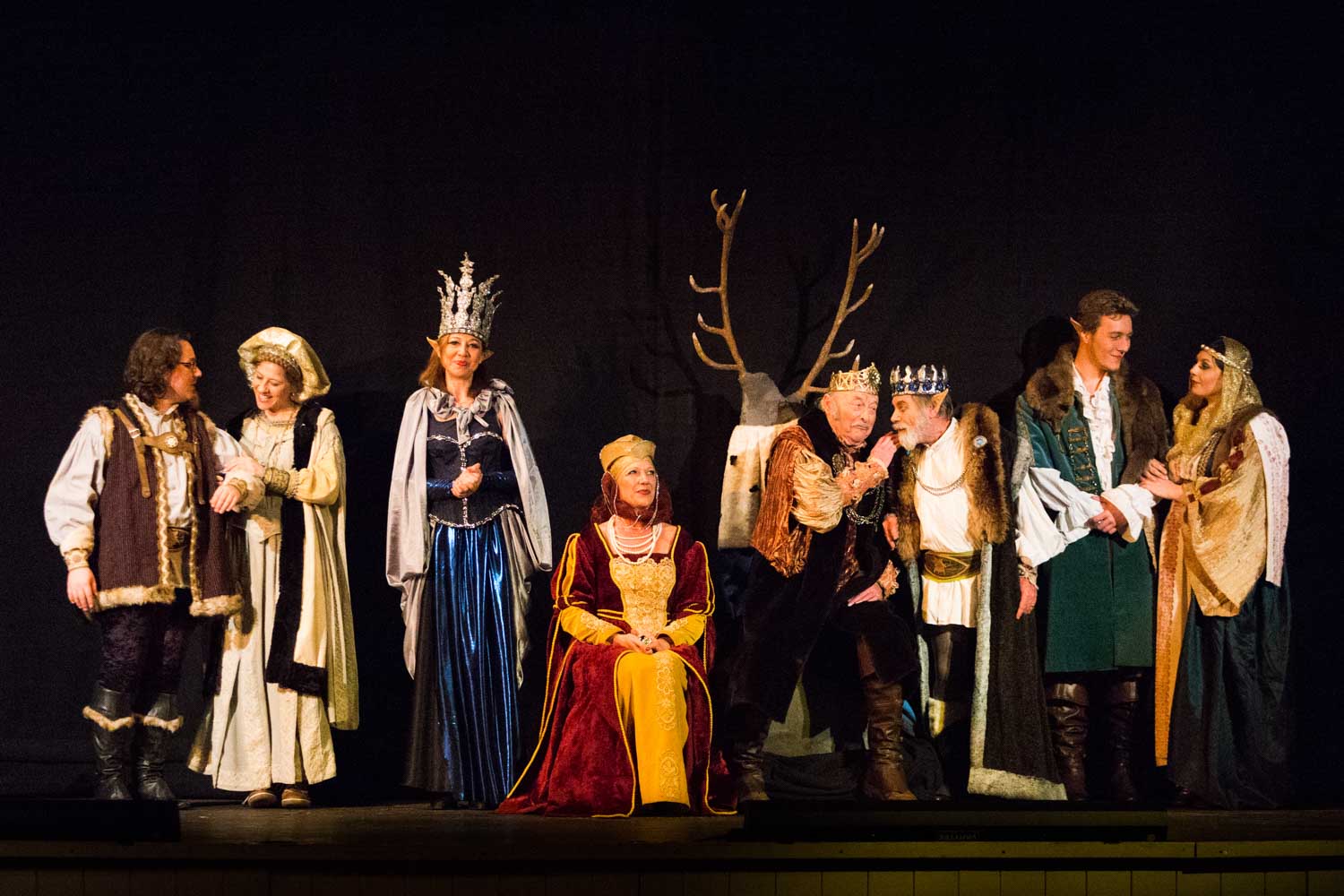 Group of actors in medieval costumes performing on stage, featuring crowns, gowns, and a backdrop with antlers.