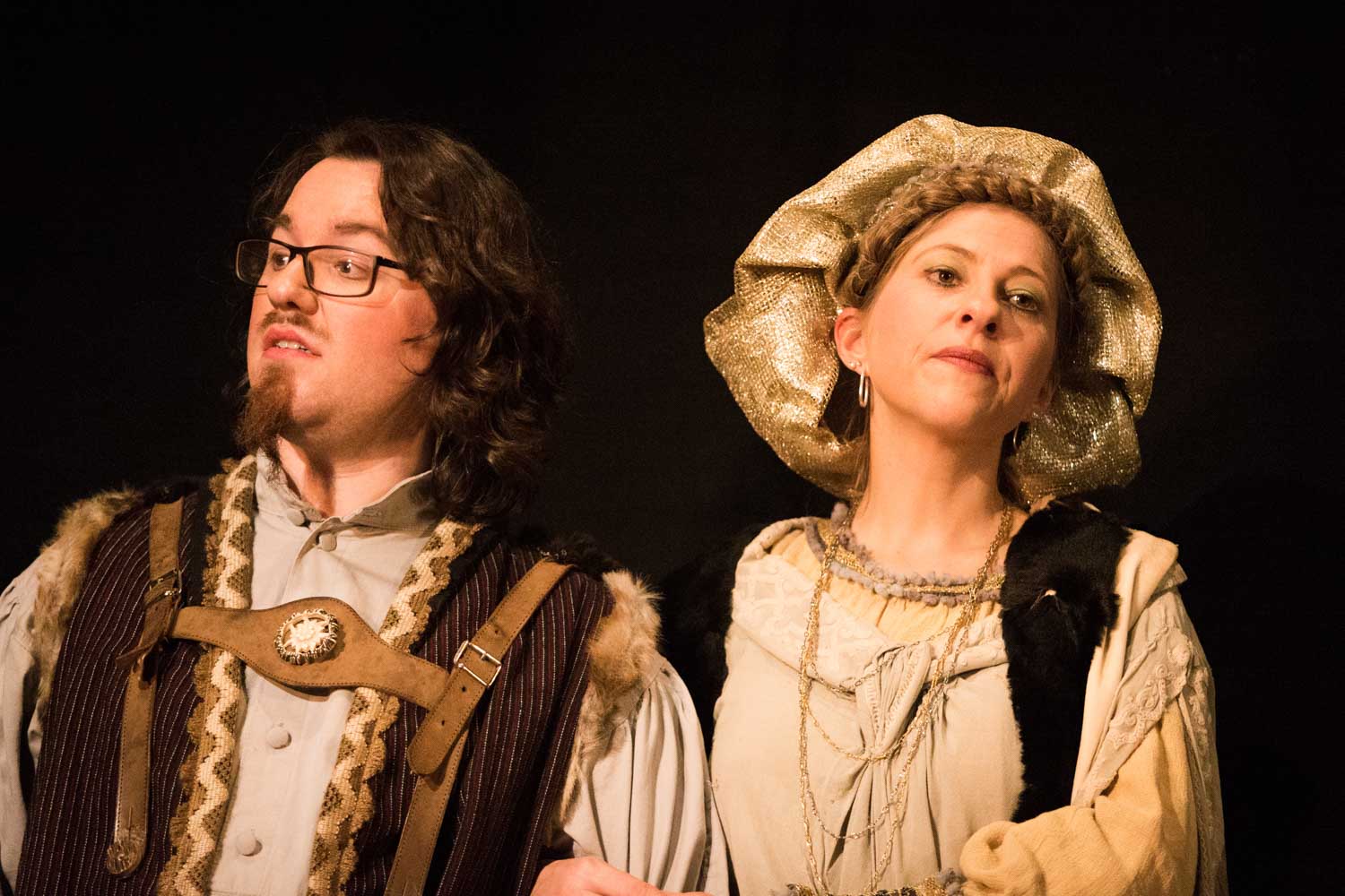Two actors in medieval costumes perform on stage, one wearing a fur-trimmed vest, the other with a gold headpiece.
