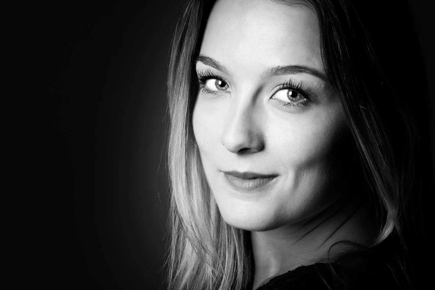 Smiling woman in black and white portrait against dark background, side-lit, showcasing detailed facial features.