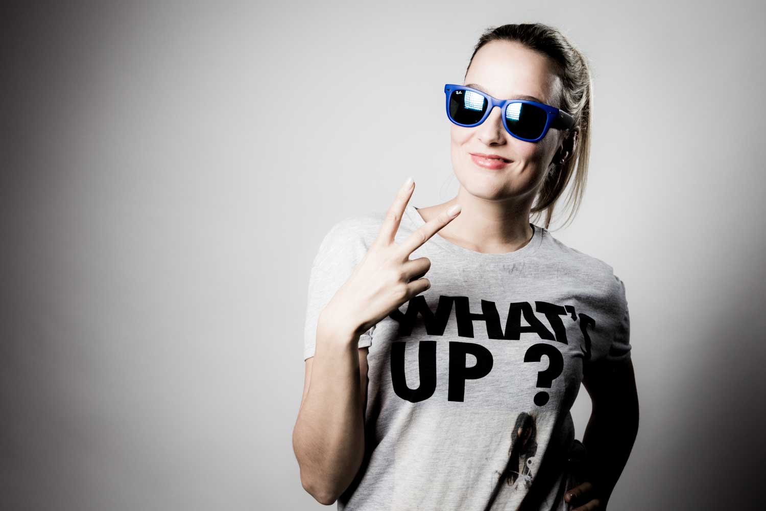 Smiling woman in sunglasses wearing a What's Up? t-shirt, making a peace sign.