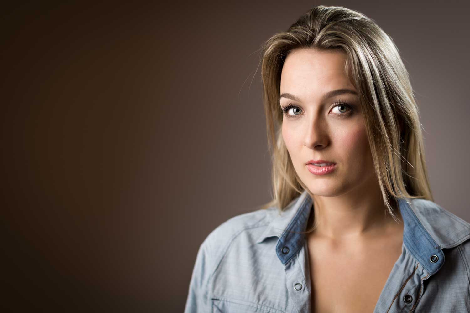 Woman with long blonde hair in a denim shirt against a brown blurred background, looking at the camera.