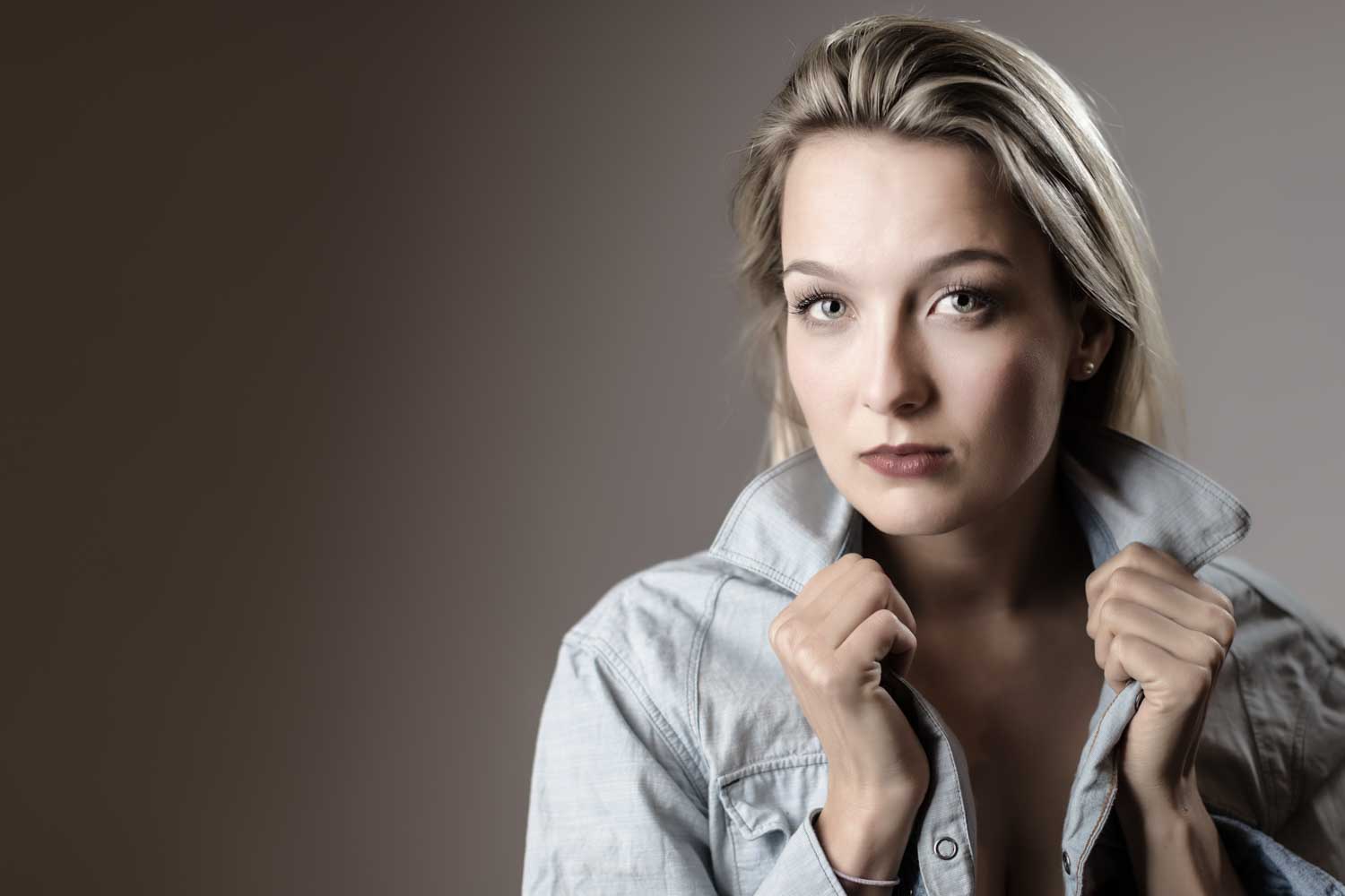 Woman in denim jacket poses with collar up, neutral background.