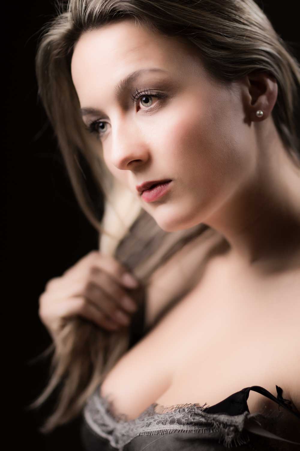 Young woman with long hair and pearl earring, wearing a lace-trimmed top, gazing thoughtfully against a dark background.
