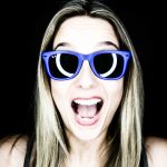 Excited woman wearing blue sunglasses, open-mouthed, against a dark background.