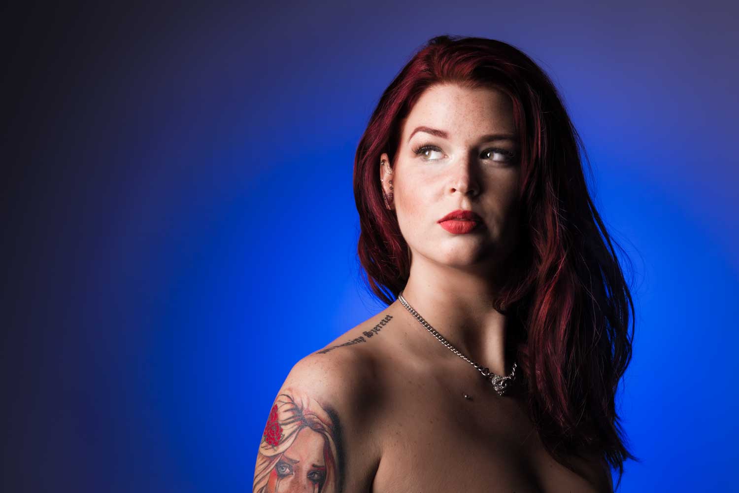 Woman with red hair and tattoo looks to the right against a blue background.