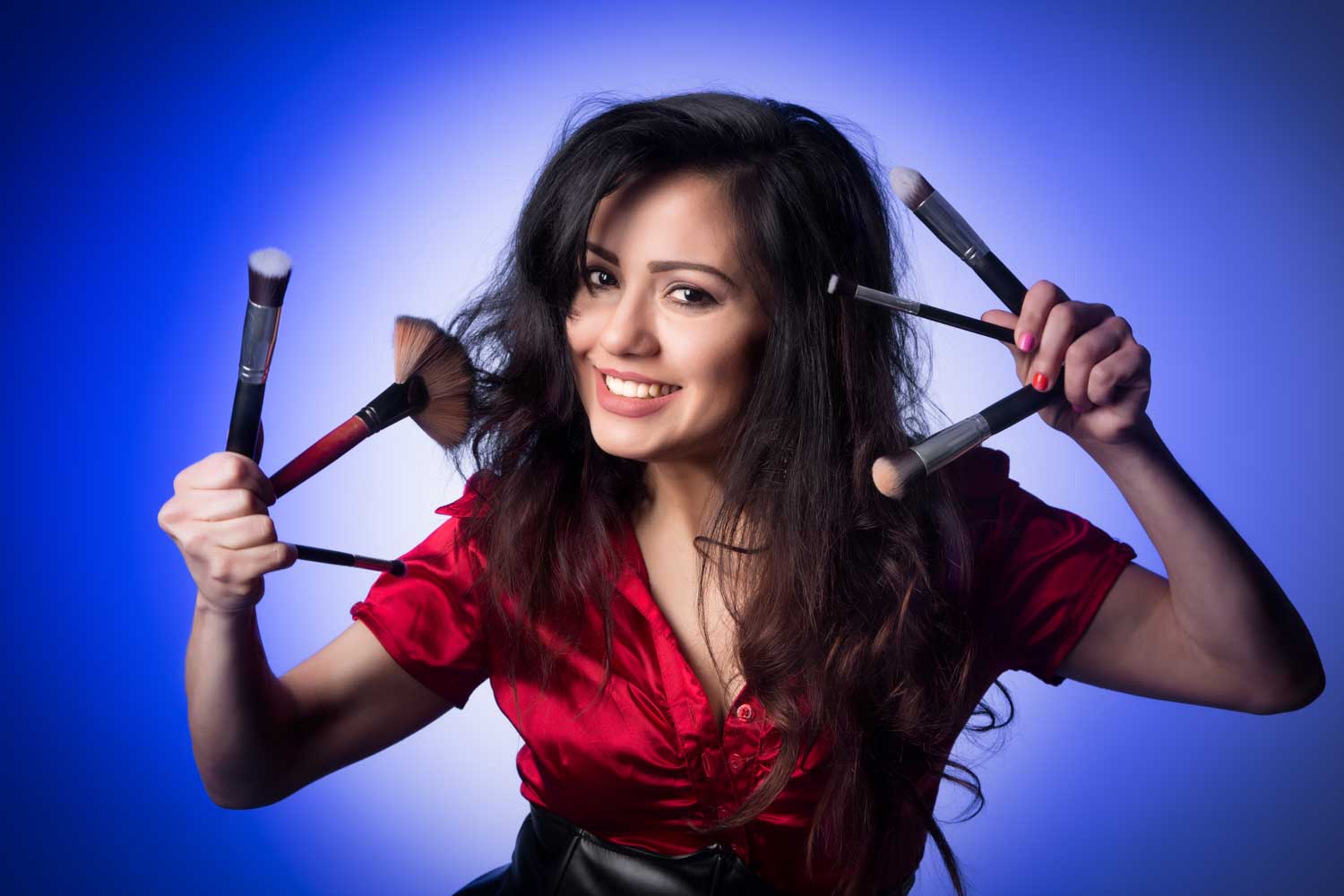 Smiling woman in red shirt holds several makeup brushes against a blue background.