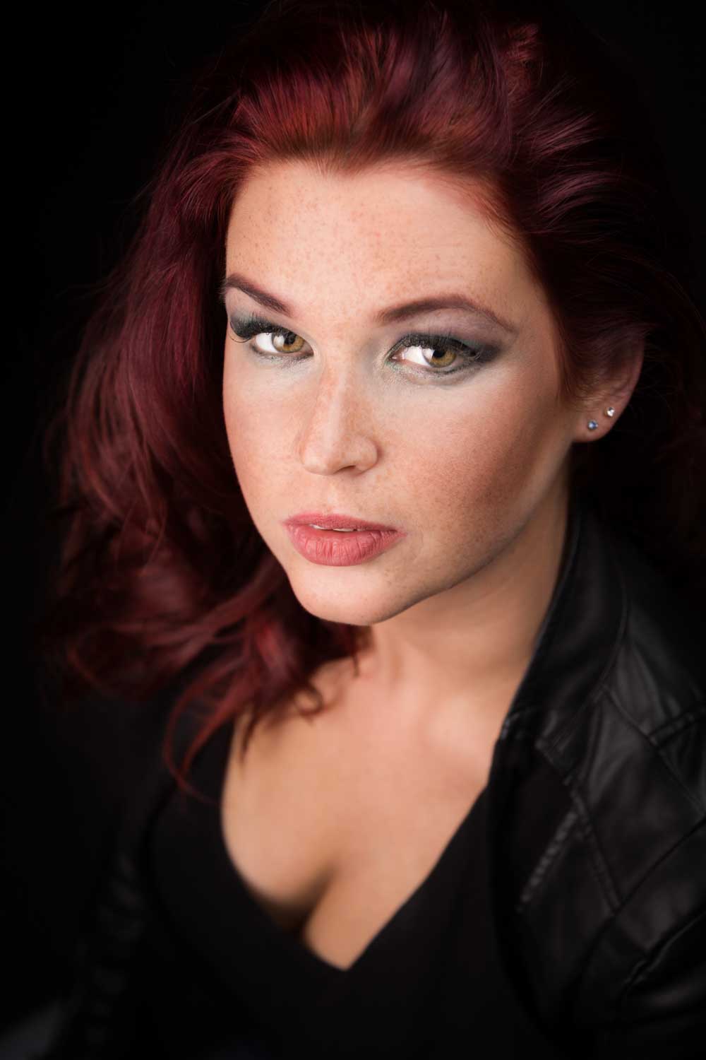 Woman with red hair and dramatic eye makeup wearing a black leather jacket against a dark background.