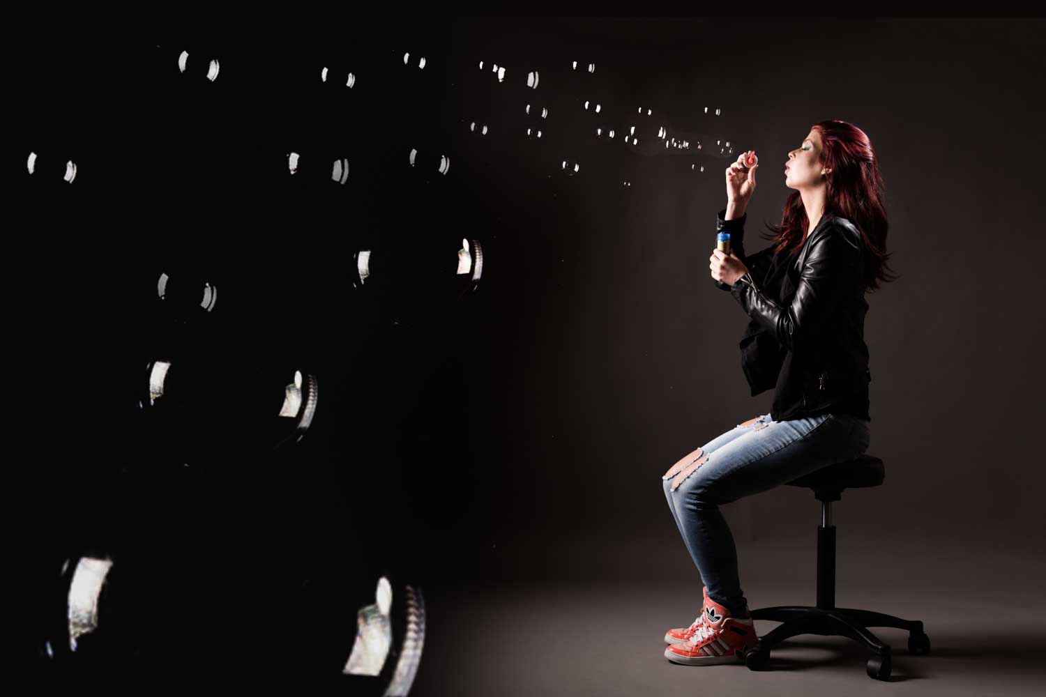 Woman in black jacket blowing bubbles while seated on a stool in a dark room.
