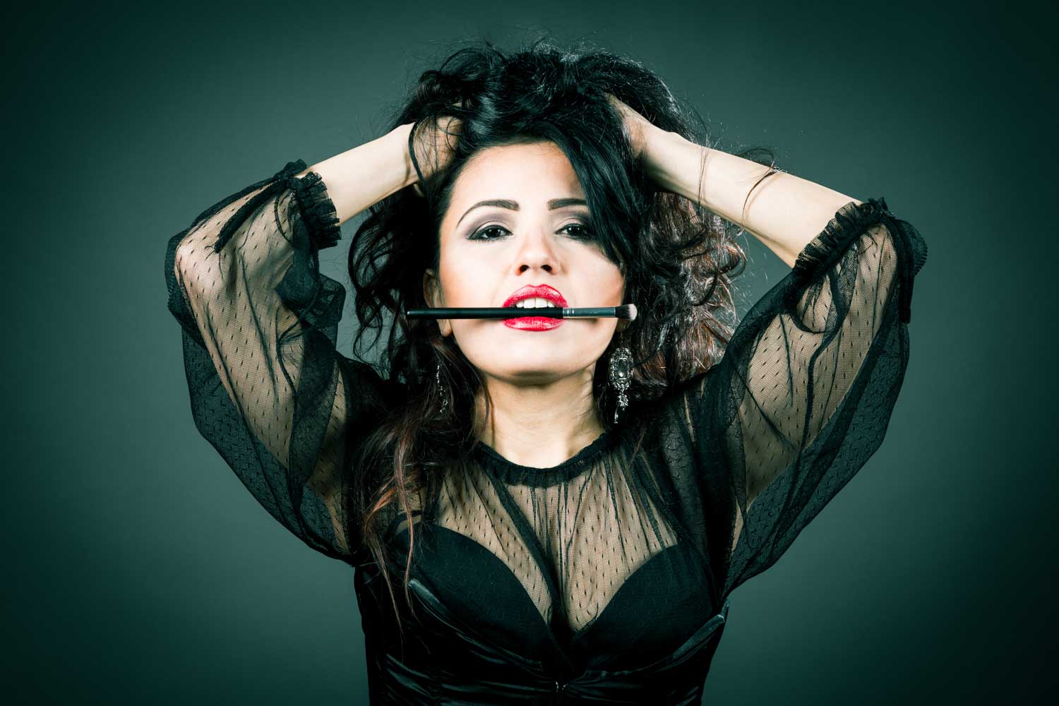 Woman with dramatic makeup holding a brush in teeth, wearing a black sheer top and holding hair with hands.