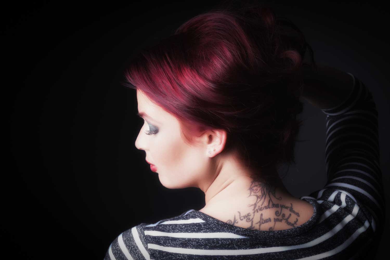 Woman with red hair and striped shirt showcasing back neck tattoo, photographed on a dark background.