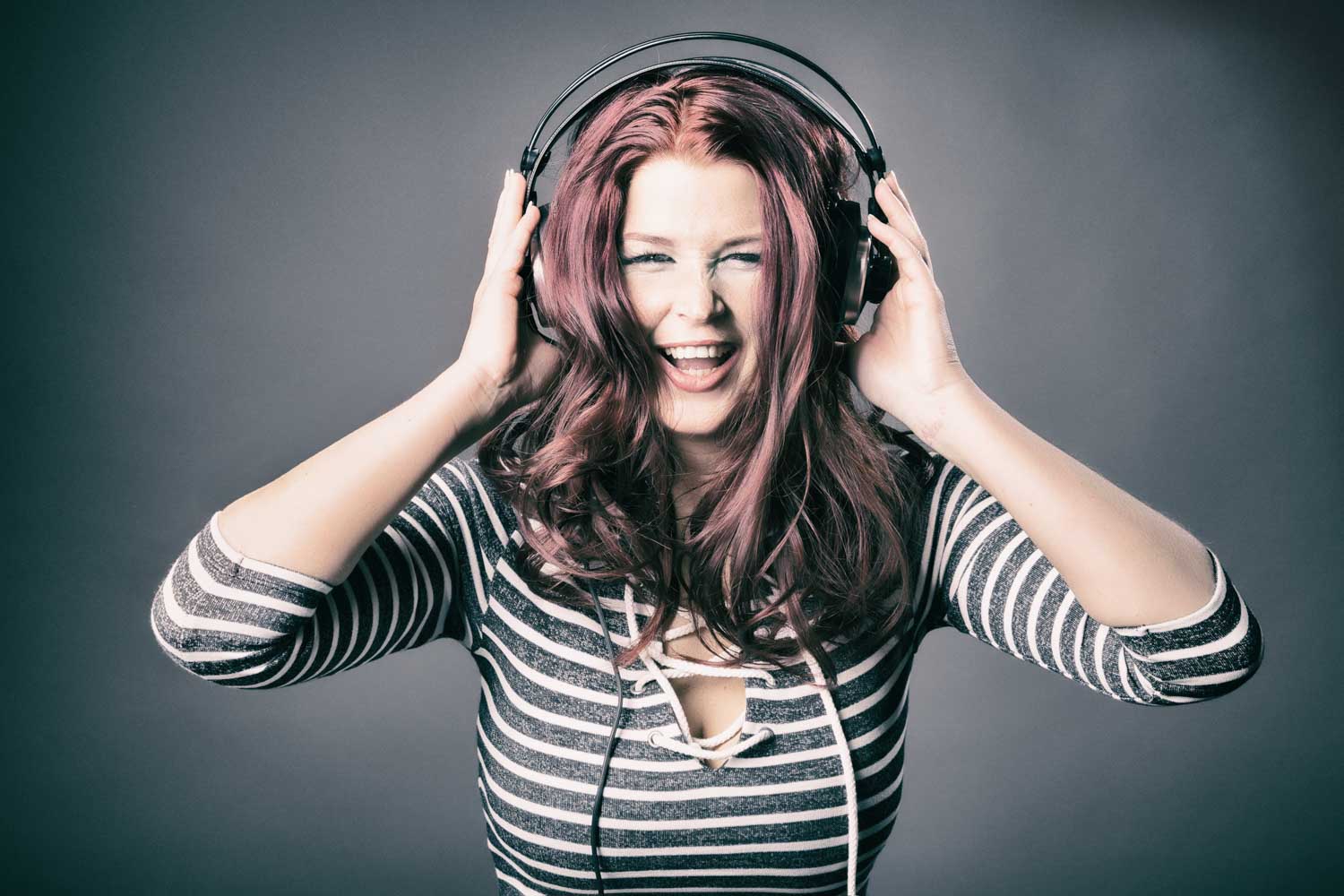 Smiling woman with red hair enjoying music on headphones, wearing a striped shirt against a gray background.