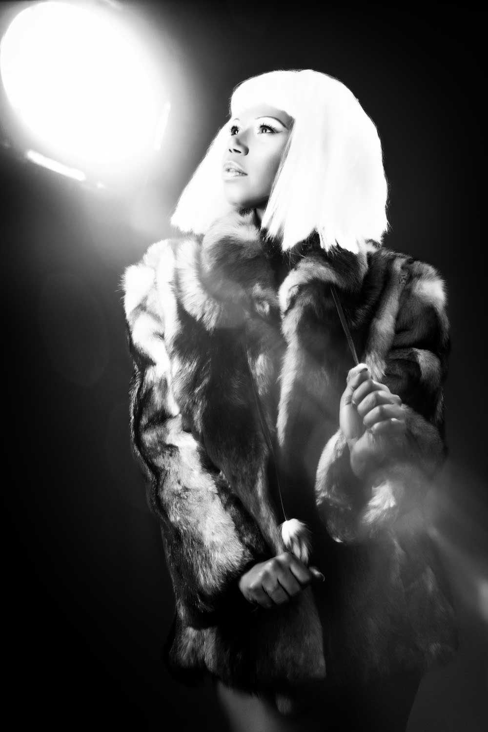 Woman in a fur coat and white wig, gazing dramatically towards a bright light in a black-and-white portrait.