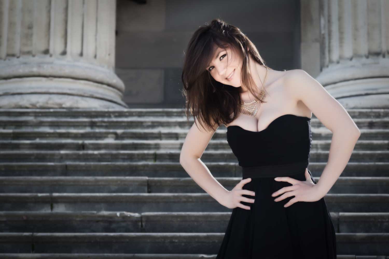 Woman in black dress posing confidently on stone steps with classical columns in background.