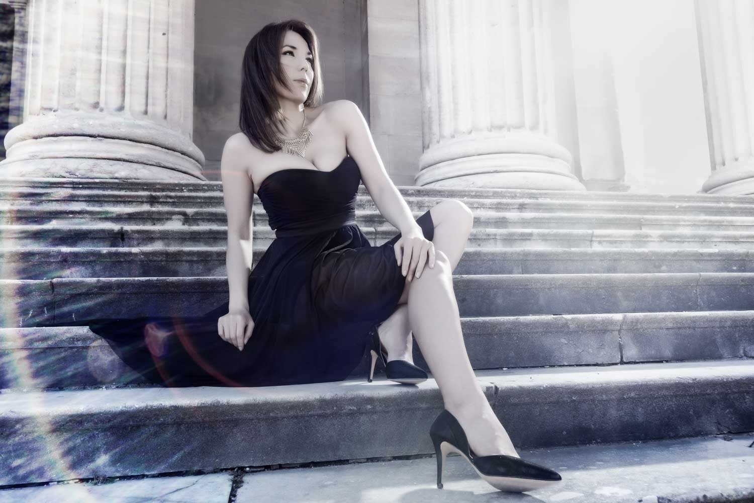 Woman in elegant black dress and heels sits on stone steps near large columns, looking away.