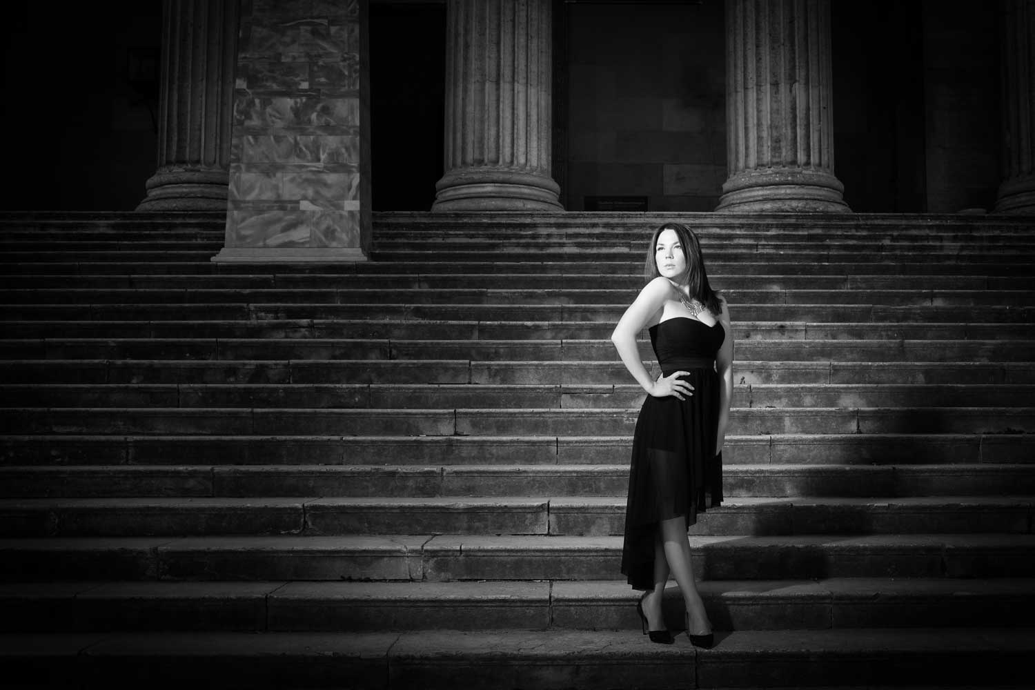 Woman in elegant black dress posing confidently on stone steps of a classical building at night.