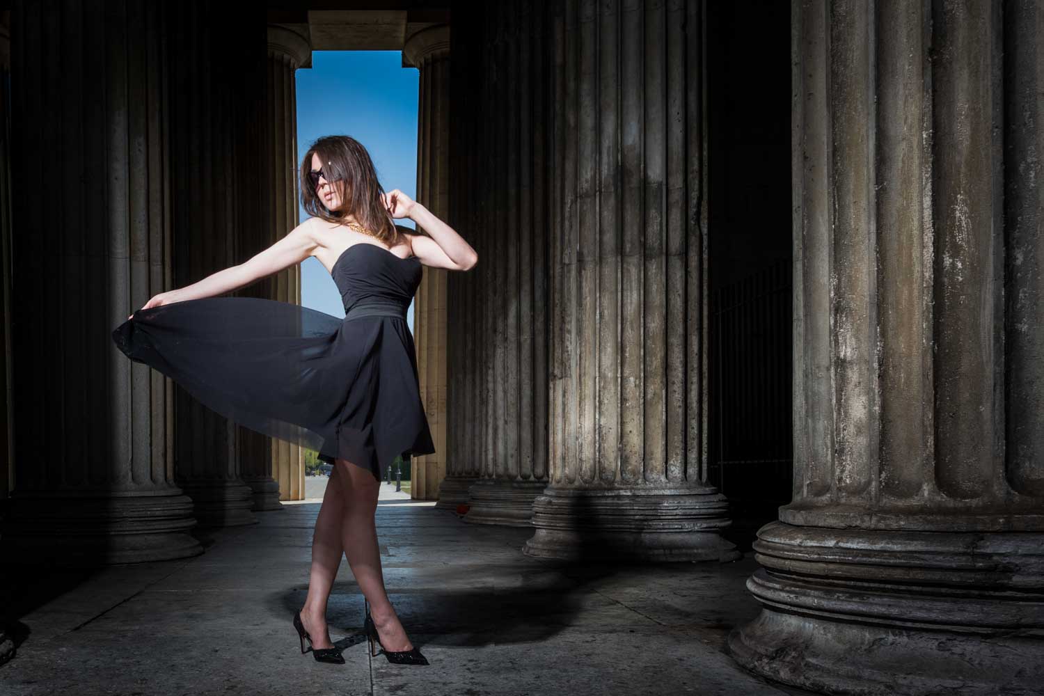 Woman in black dress posing between large stone columns, sunlight casting shadows, creating a dramatic and stylish effect.