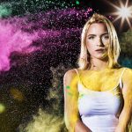 Blonde woman in white tank top with colorful powder on face and body, vibrant pink and green powder in the air.