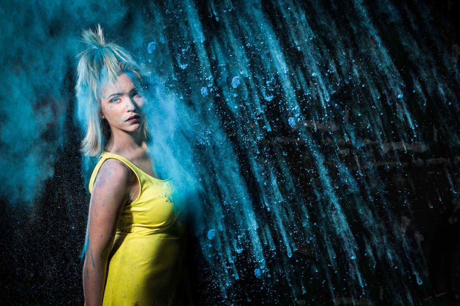 Woman in yellow dress amidst vibrant blue powder splash, creating dynamic motion against dark background.