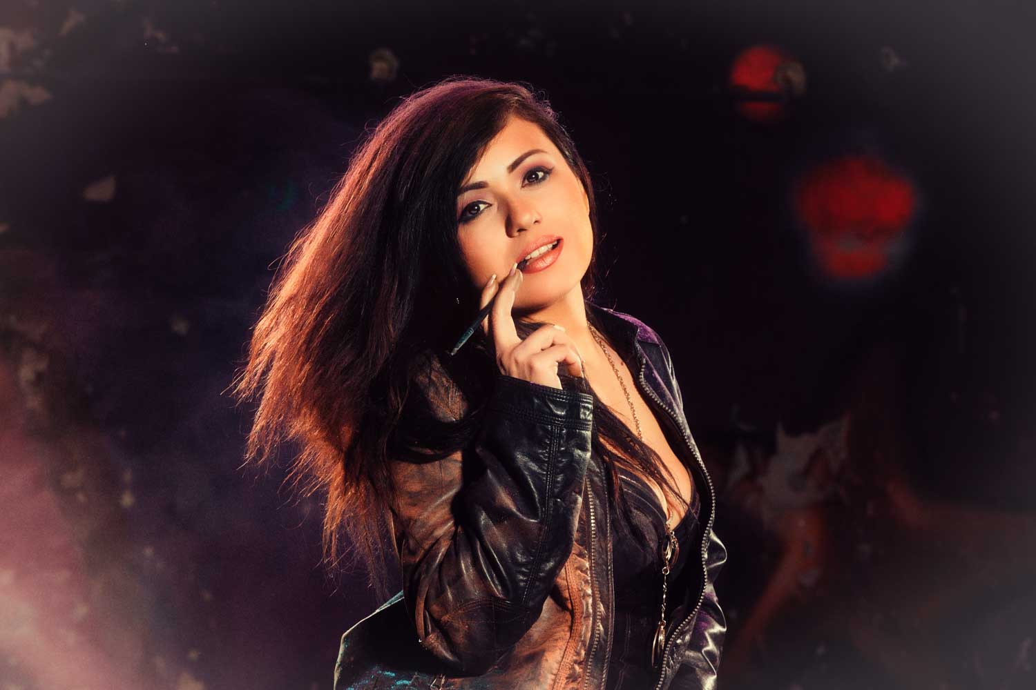 Woman in leather jacket poses confidently, holding a pen to her lips against a dark, atmospheric background.