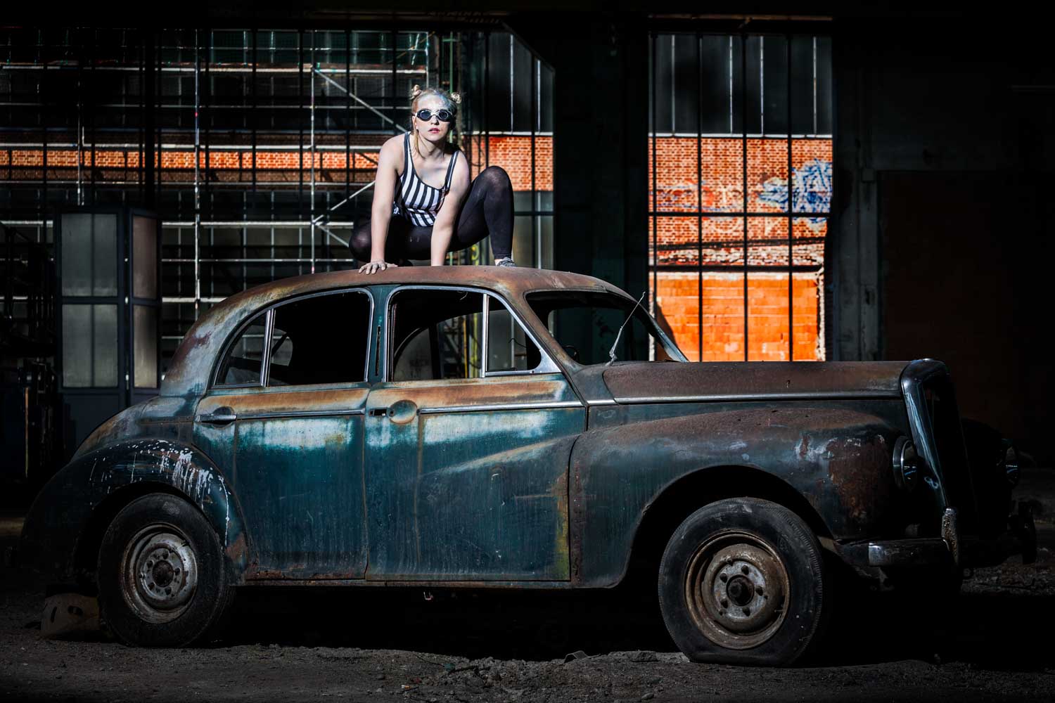 Woman in striped outfit crouches on rusty vintage car in an industrial warehouse.