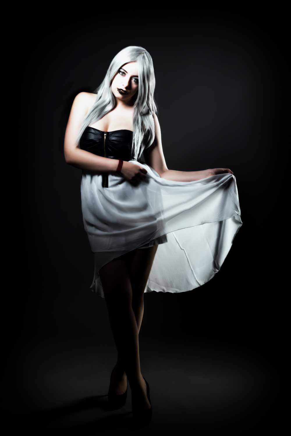 Woman in black top and white skirt with long silver hair poses dramatically against a dark background.