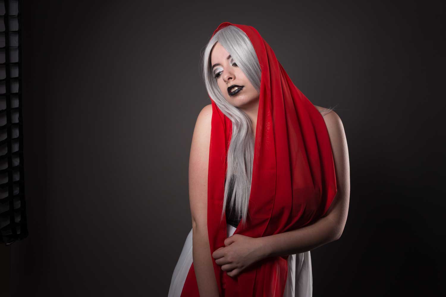 Woman with long silver hair, black lipstick, and red shawl posing against dark backdrop.