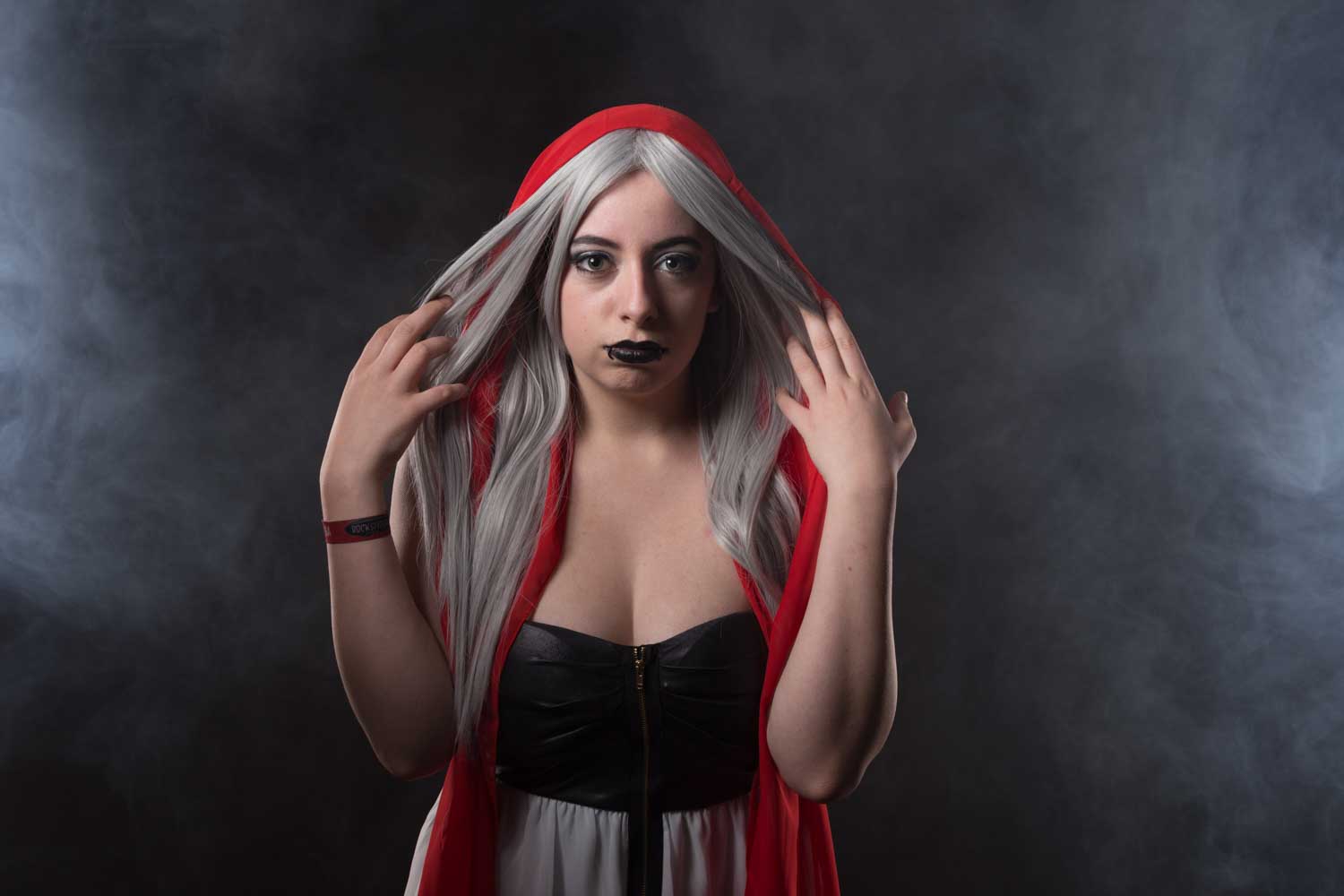 Woman in a red hood with silver hair and black lipstick against a smoky background.