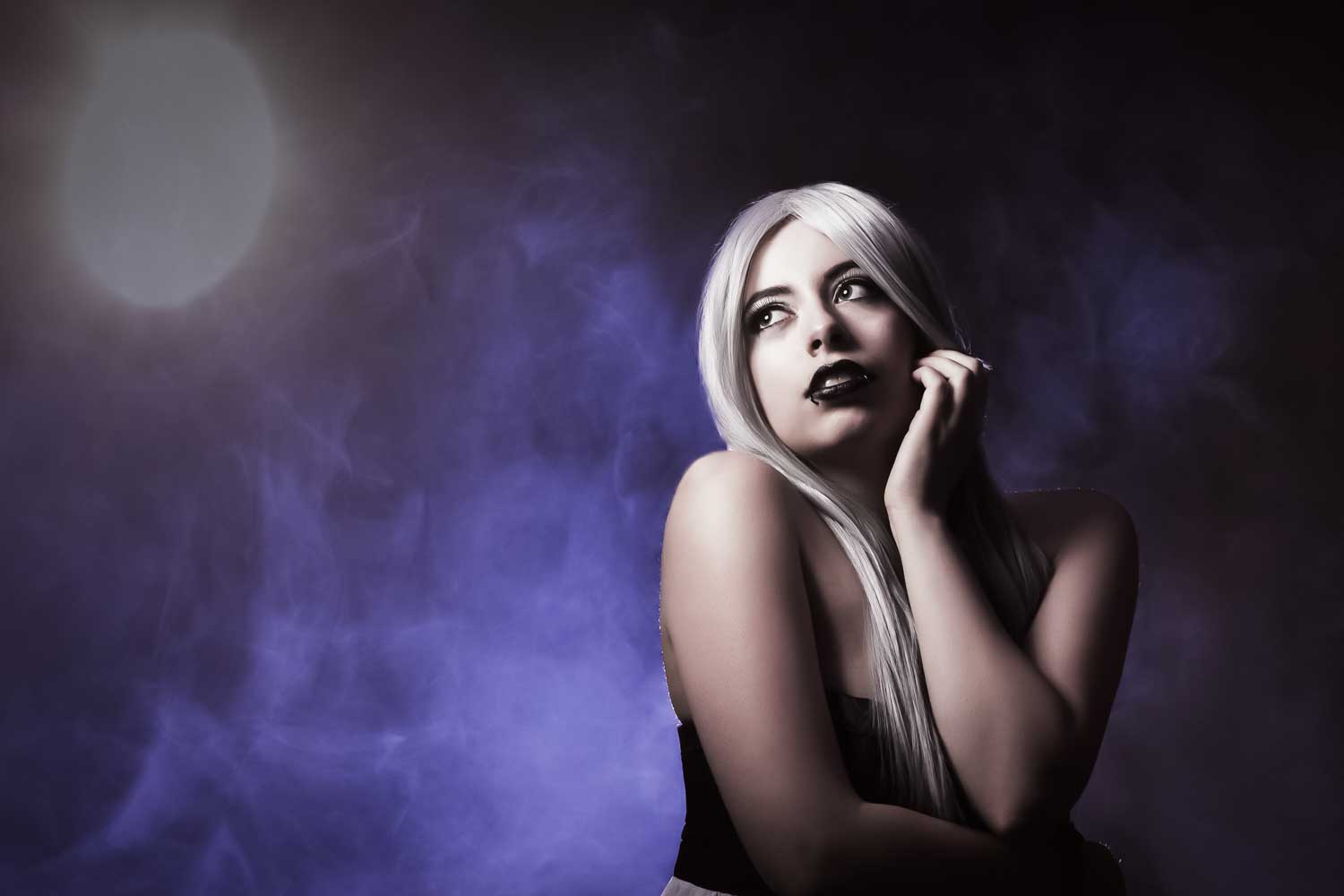 Woman with long white hair and black lipstick gazes thoughtfully against a misty purple background.