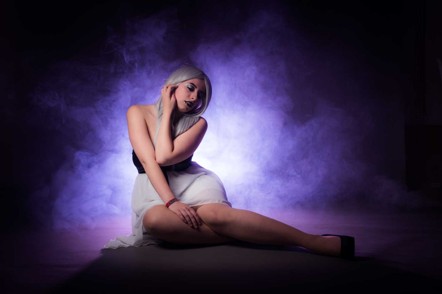 Model in gray wig and white dress posing against a purple smoke background in a moody, artistic setting.