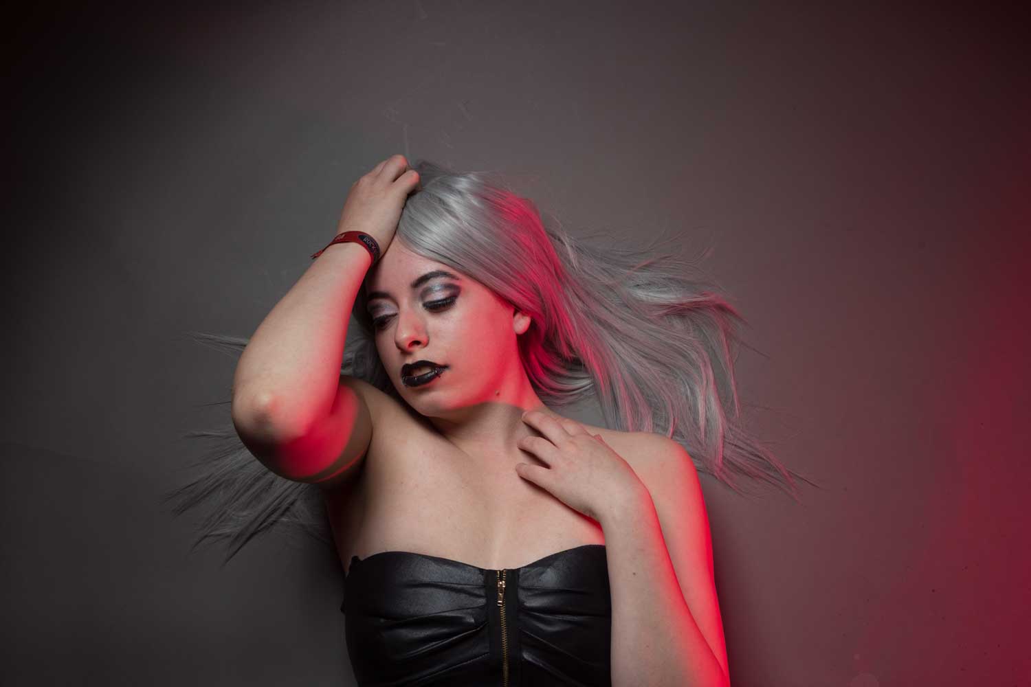 Woman with silver hair, black lipstick, and leather top lying down; accented by dramatic red lighting.