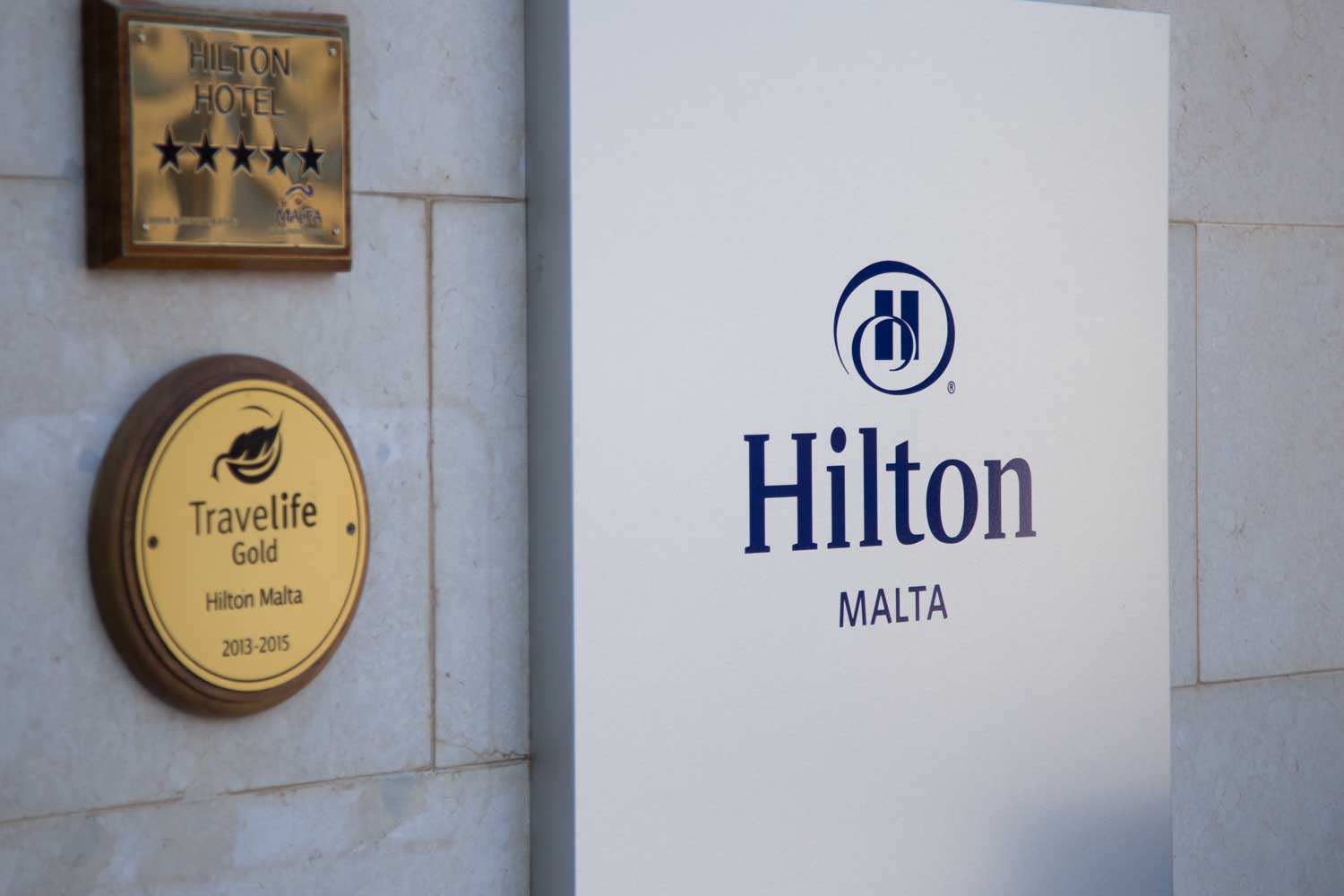 Hilton Malta hotel sign with five-star and Travelife Gold awards displayed on a light stone wall.