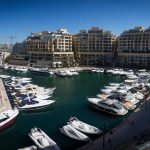 Luxury yachts docked in a marina surrounded by modern buildings under a clear blue sky.