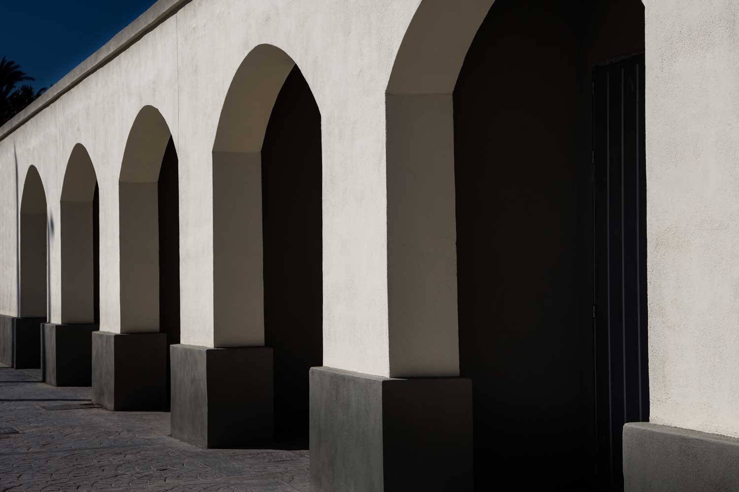 Arched walkway with contrasting shadows against a light stucco wall, emphasizing architectural symmetry and texture.