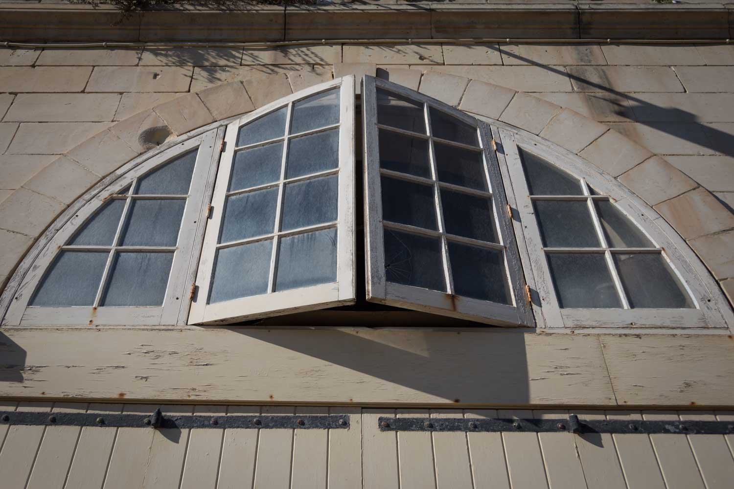 Arched wooden window with peeling paint partially open against a stone wall, casting a shadow in sunlight.