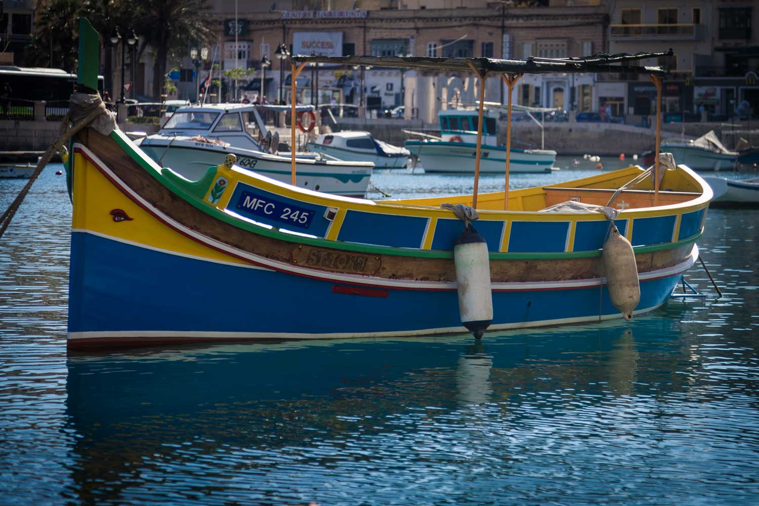 Colorful traditional wooden boat moored in a harbor, reflecting on calm blue water with buildings in the background.